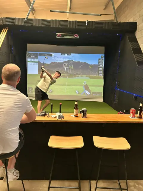 A golfer mid-swing in a simulator bay while a friend watches from the bar ledge with drinks